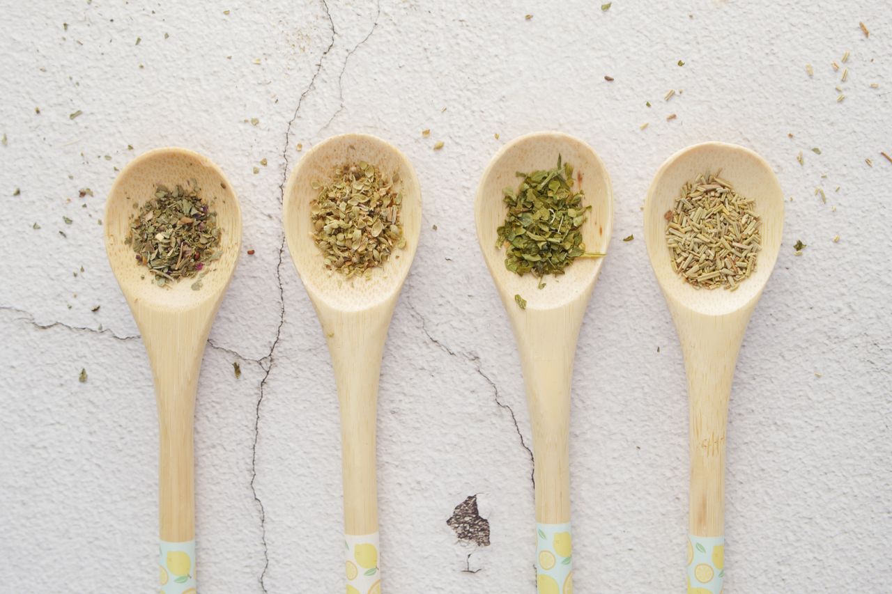 Four wooden spoons with assorted dried herbs on a light background.